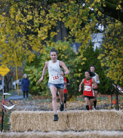 Lauren, a Rose City Track Club member, jumping a hay hurdle during a track event with competitors in the distance.